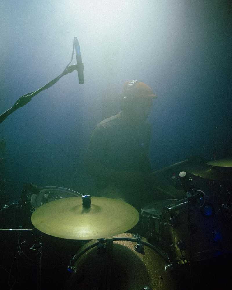 A man behind a drumkit, in fog wearing a flatcap